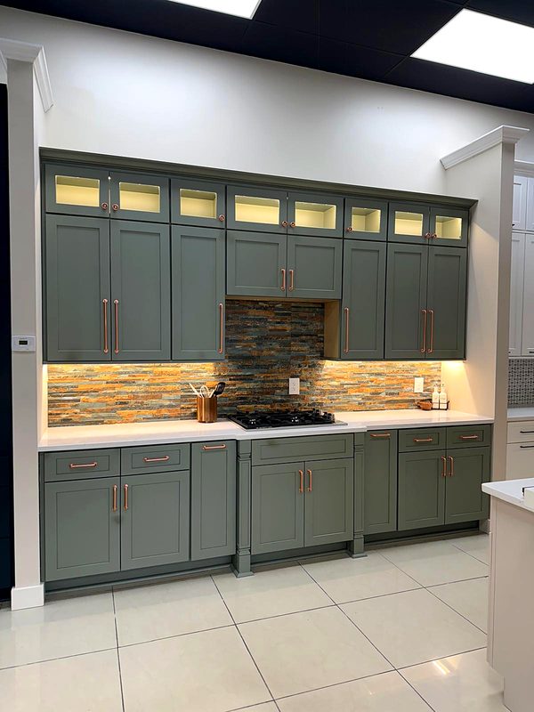 Sage/olive green shaker cabinets with copper pulls, lighted glass-top display cubbies — on display at MNK Cabinet's Norcross, GA showroom