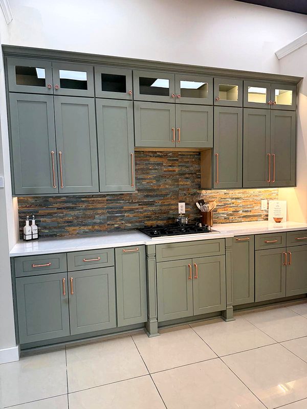 Sage green shaker cabinets with copper/rose-gold pulls and knobs, lighted glass-top — kitchen by MNK Cabinet in metro Atlanta
